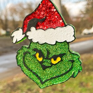 May include: A glittery green Grinch face ornament with a red glitter Santa hat and white trim. The Grinch's eyes are yellow, and the outline is black. The ornament hangs from a black string and silver ring.