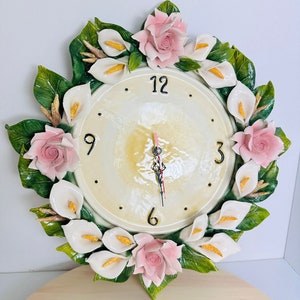 May include: A white and green ceramic wall clock with a floral design. The clock face is white with black numbers and hands. The flowers are pink and white and are arranged in a circle around the clock face.