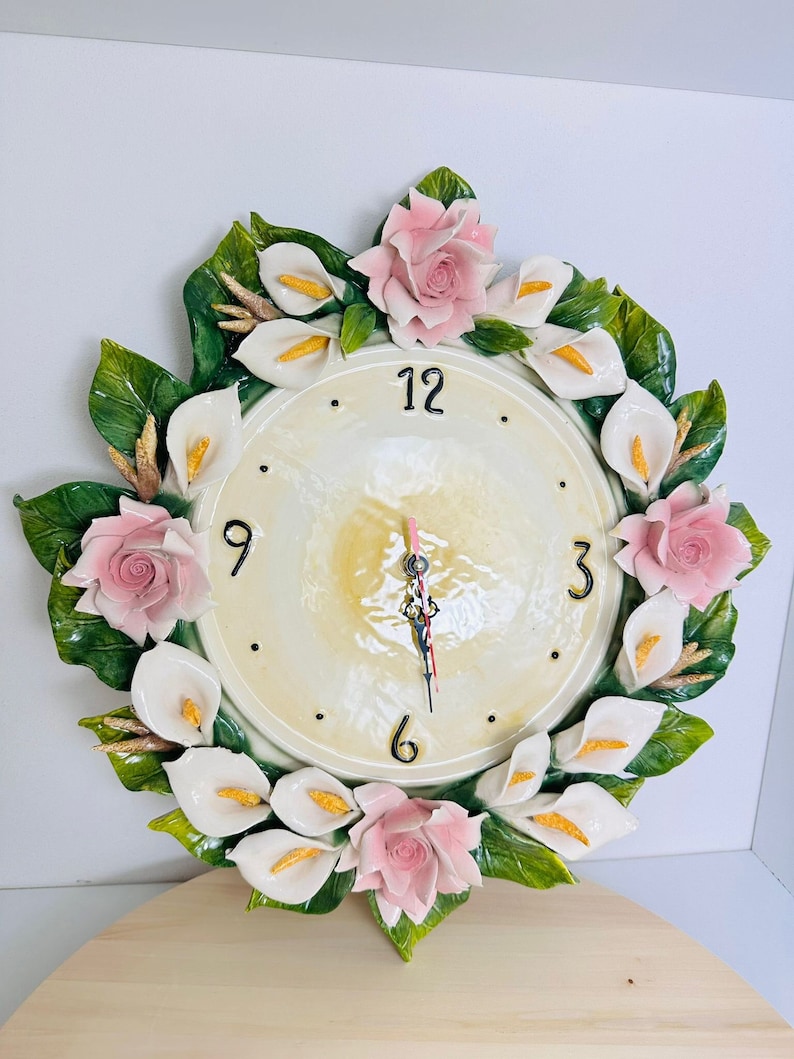 May include: A white and green ceramic wall clock with a floral design. The clock face is white with black numbers and hands. The flowers are pink and white and are arranged in a circle around the clock face.