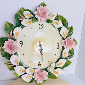 May include: A white and cream floral clock with pink roses and white calla lilies. The clock face has black Roman numerals and black hands.