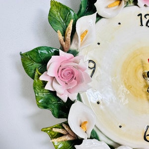 May include: A white ceramic clock with a floral design. The clock face has Roman numerals and is surrounded by white calla lilies, pink roses, and green leaves.