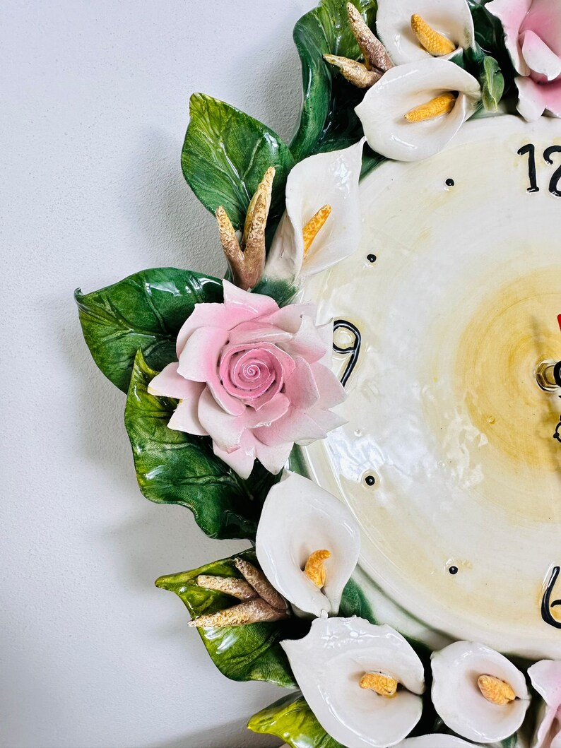 May include: A white ceramic clock with a floral design. The clock face has Roman numerals and is surrounded by white calla lilies, pink roses, and green leaves.