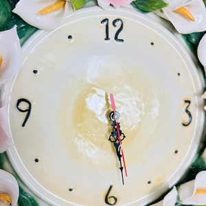 May include: A white ceramic clock with a floral design. The clock face is white with black numbers and hands. The floral design features white calla lilies, pink roses, and green leaves.