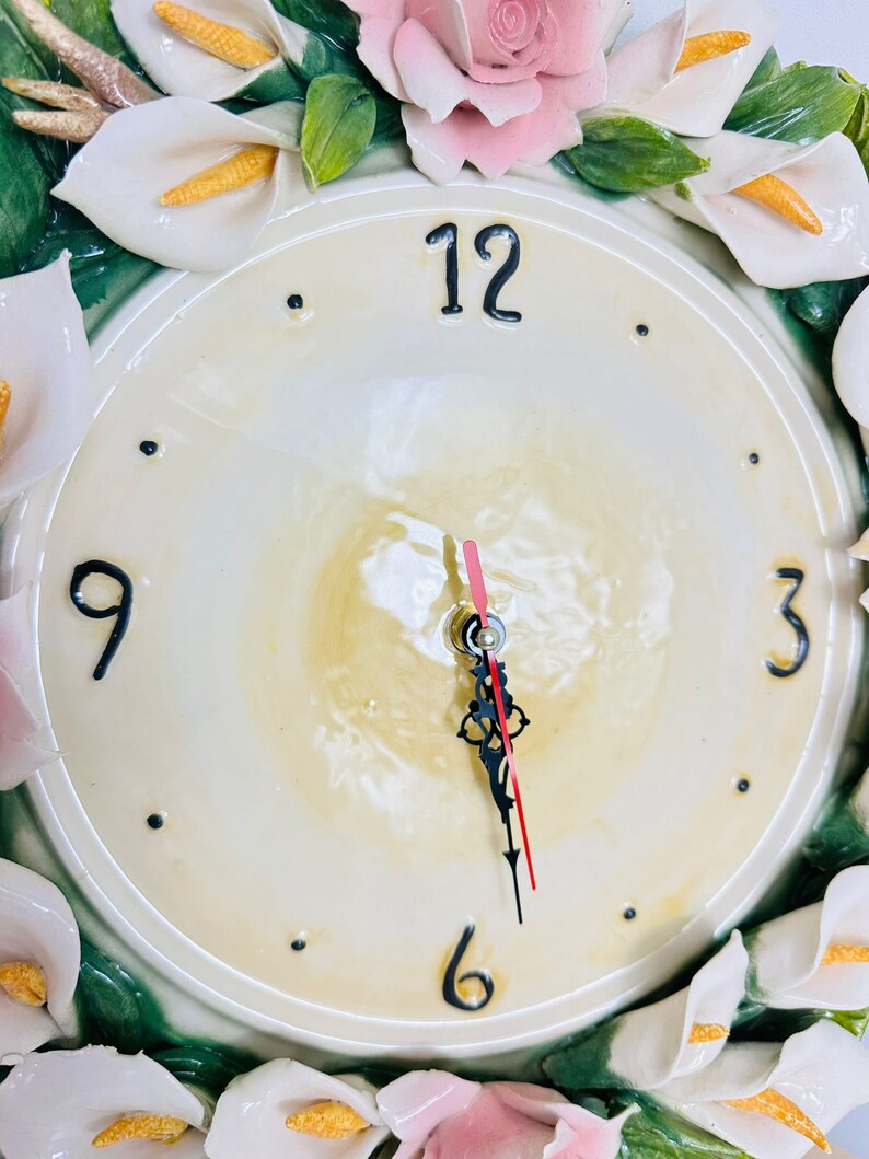 May include: A white ceramic clock with a floral design. The clock face is white with black numbers and hands. The floral design features white calla lilies, pink roses, and green leaves.