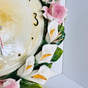May include: A white ceramic clock face with black Roman numeral 3, surrounded by a wreath of white calla lilies and pink roses with green leaves.