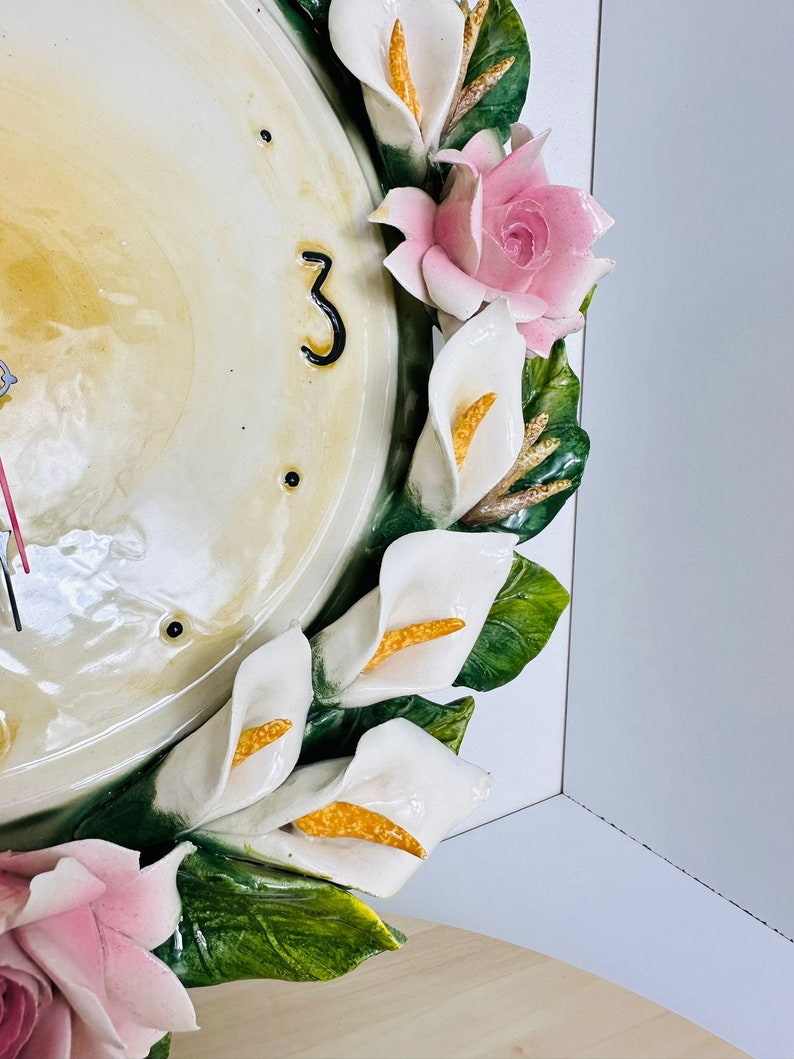 May include: A white ceramic clock face with black Roman numeral 3, surrounded by a wreath of white calla lilies and pink roses with green leaves.