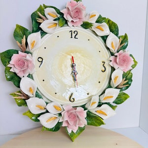 May include: A white ceramic clock with a floral design. The clock face is white with black numbers and hands. The floral design features pink roses and white calla lilies.