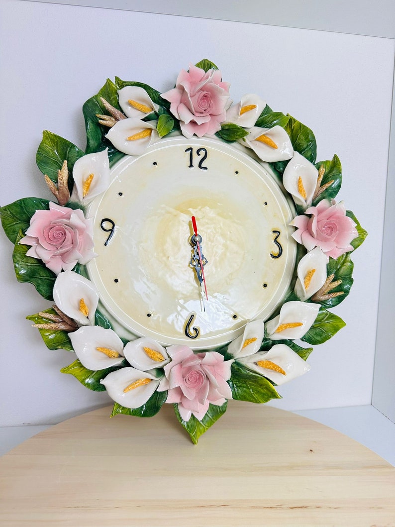 May include: A white ceramic clock with a floral design. The clock face is white with black numbers and hands. The floral design features pink roses and white calla lilies.
