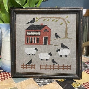 May include: A framed cross-stitch design depicting a red building with the text "Black Crow Woolen Mill" and a flock of sheep in front of it. The design is set against a grey background with a black fence and a tree with green leaves.