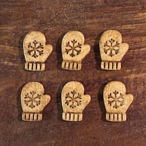 May include: Six wooden mitten-shaped buttons with a snowflake design. The buttons are light brown with a darker brown snowflake and ribbed cuff detail. The buttons are arranged on a dark brown wooden surface.
