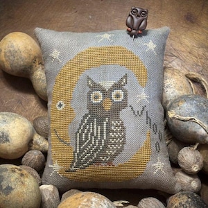 May include: A square gray fabric pincushion featuring a cross-stitched design of a dark brown owl perched on a golden crescent moon with small white stars. The word whoo is stitched in black script. A small brown owl-shaped pin is attached.