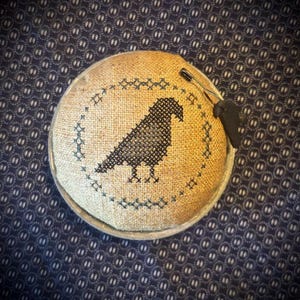 May include: A round, tan linen pincushion featuring a dark blue cross-stitched raven silhouette centered within a circular border of small cross-stitch patterns. A silver-toned safety pin with a small black charm is attached to the top edge.