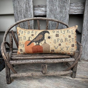 May include: A rectangular, beige cross-stitch pillow featuring a crow holding a spider, perched above three pumpkins. The pillow has an alphabet and decorative symbols. The pillow is on a small, rustic wooden bench.
