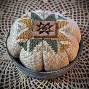 May include: A round, stuffed fabric pincushion featuring a cross-stitched star pattern in shades of olive green and mustard yellow on a beige linen background. A square, carved wooden button with a star cutout sits in the center of the cushion.