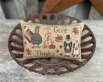 May include: A beige pillow with a cross-stitch design featuring a turkey, pumpkins, corn stalks, and two figures in pilgrim clothing. The text "Give Thanks" is stitched on the pillow.