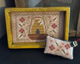 May include: A framed cross-stitch featuring a beehive, bees, and flowers in shades of brown, yellow, and red. A small, matching pin cushion with a button and pin is next to the framed art. The frame is yellow.