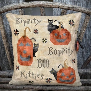 Peut inclure: Un coussin beige brodé avec des motifs d'Halloween. Il présente des citrouilles oranges, des chats noirs et les mots "Rippity", "Boppity", "Boo" et "Kitties". Des éléments floraux décoratifs sont également présents.