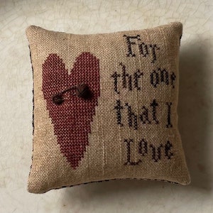 Puede incluir: Una peque&ntilde;a almohada de arpillera marr&oacute;n con un coraz&oacute;n rojo bordado en la parte delantera. El coraz&oacute;n tiene un imperdible atravesado y las palabras "Para el que amo" est&aacute;n bordadas en negro en la almohada.