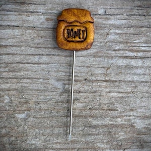 May include: A brown, textured, jar-shaped pin with a lid and the word HONEY embossed on the front. The pin features a long, thin, silver-colored metal needle extending from the bottom of the jar shape, designed for securing or decorative purposes.