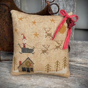 May include: A beige, square decorative pillow with a cross-stitched Christmas scene. The design features Santa in a sleigh pulled by reindeer, a house, trees, and stars. A red ribbon and sleigh charm are attached to the pillow.