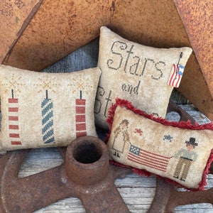 May include: Three embroidered pillow covers with a patriotic theme. The first pillow features a red, white, and blue firework design. The second pillow says "Stars and Stripes" in a brown font. The third pillow features a red, white, and blue flag and two figures in traditional clothing.