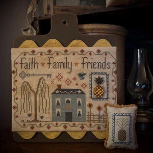 May include: A framed cross-stitch sampler with the words "Faith Family Friends" and a house, trees, and pineapple design. A small pillow with a pineapple design sits in front of the sampler. The frame is brown with a yellow scalloped edge.