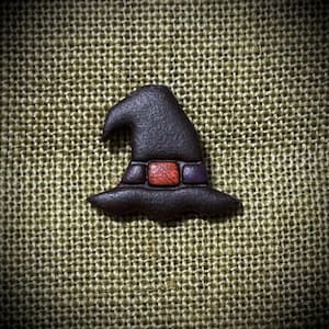 May include: A dark brown witch hat pin with a textured surface. The hat features a wide brim and a pointed top that curves slightly to the left. A rectangular band sits at the base of the crown, colored in shades of orange and purple.