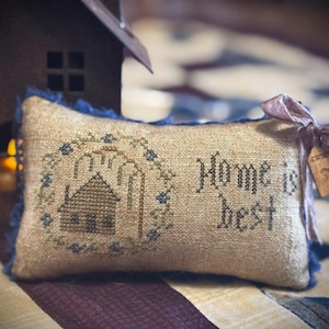 Könnte beinhalten: Ein rechteckiges, beiges Kreuzstichkissen mit den Worten "Home is best" und einem Hausdesign. Das Kissen hat blaue Fransen und ein rosa Band mit einem kleinen Anhänger. Ein Miniaturhaus befindet sich im Hintergrund.