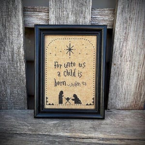 May include: A framed cross-stitch artwork in a black frame. The artwork features the text "For unto us a child is born...Isaiah 9:6" and a star above a silhouette of a nativity scene. The background is tan.