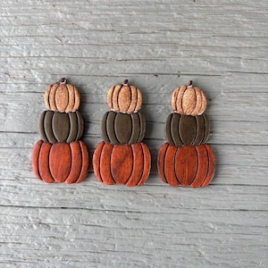 May include: Three stacked wooden pumpkins in shades of orange, brown, and tan. Each pumpkin stack has three sections, with the largest at the bottom and the smallest at the top. The pumpkins are arranged on a weathered, gray wooden surface.