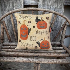 May include: A beige, square decorative pillow with a Halloween theme. The pillow features cross-stitch designs of orange jack-o'-lanterns, black cats, and the words "Bippity Boppity Boo Kitties". The pillow is resting on a rustic wooden chair.