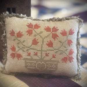 May include: A small, rectangular pillow with a beige fabric base. The pillow features a cross-stitched floral design in shades of red and green. The edges are trimmed with a fringed border in a matching beige color. The pillow is likely handmade.