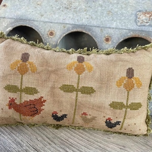 Puede incluir: Una almohada beige con un borde de flecos verdes presenta un diseño bordado de tres flores amarillas con centros marrones, un gallo rojo y tres pollos negros.