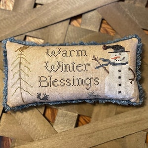 Peut inclure: Un coussin beige avec une frange bleue présente un motif de point de croix représentant un bonhomme de neige avec un chapeau noir et une écharpe, un pin et le texte "Warm Winter Blessings".