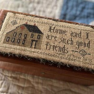 Peut inclure: Une boîte en bois avec un dessus en tissu cousu à la main. Le tissu présente une petite maison avec un toit rouge et le texte "Home and I are such good friends".