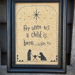 May include: Framed cross-stitch artwork with a black frame. The artwork features the text "For unto us a child is born...Isaiah 9:6" in black thread. A star and silhouettes of figures are also stitched.