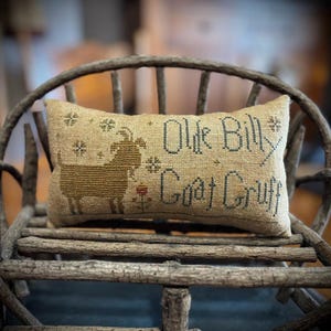 Könnte beinhalten: Ein rechteckiges beiges Leinenkissen mit einer im Kreuzstich gestickten braunen Ziege, einer kleinen Blume und dem Text "Olde Billy Goat Gruff" in dunkelblauem Garn. Das Kissen ist mit kleinen sternförmigen Akzenten verziert und hat eine Textur.