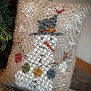 Peut inclure: Un coussin décoratif brodé au point de croix avec un bonhomme de neige. Le bonhomme de neige porte un chapeau noir avec une étoile dorée et une écharpe rouge. Il a un nez en carotte et est orné de boules colorées. Des flocons de neige et un oiseau rouge sont également brodés sur le tissu beige.