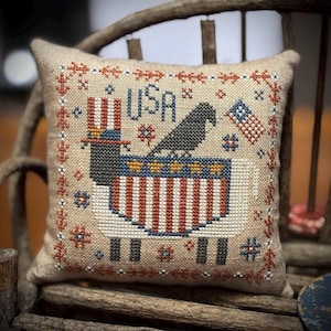 May include: A square throw pillow made of tan linen fabric featuring a cross-stitch design of a sheep wearing a top hat with red and white stripes. The sheep has a blue body with white stars and red and white stripes. The text USA is stitched in blue.