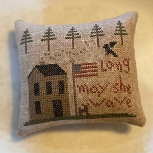 May include: A beige pillow with a cross-stitch design featuring a house, trees, a flag, and the text "May the long wave".