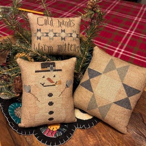 Peut inclure: Trois petits coussins en toile de jute avec des motifs brodés. Un coussin représente un bonhomme de neige avec un chapeau haut de forme noir, un autre a un motif d'étoile dans les tons bleus et gris, et le troisième coussin porte le texte "Mains froides, mitaines chaudes".