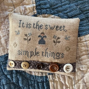 May include: A beige pillow with the text "It is the sweet simple things" embroidered on it. The pillow has a brown trim with five buttons.