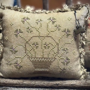 May include: A square beige fabric pillow featuring a cross-stitched design of a vase filled with flowers in muted green and purple tones. The pillow is finished with a frayed fabric edge and includes a small metal clover charm attached to the side.
