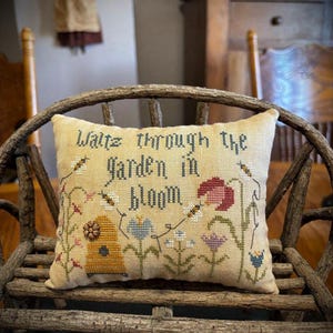 May include: A decorative, rectangular pillow with the embroidered text "Waltz through the Garden in Bloom." The pillow features a garden scene with flowers, bees, and a beehive. The pillow is placed on a rustic wooden chair.