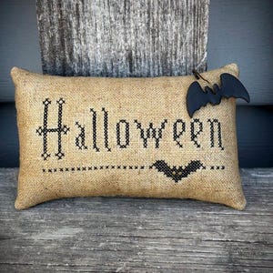 Peut inclure: Un coussin rectangulaire en toile de jute avec le mot "Halloween" brodé en fil noir. Une décoration en forme de chauve-souris noire est attachée. Le coussin repose sur une surface en bois vieilli.