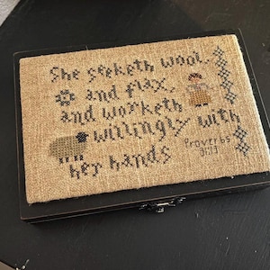 May include: A framed cross-stitch sampler with the text "She seeketh wool, and flax, and worketh willingly with her hands" and the Bible verse "Proverbs 31:13".