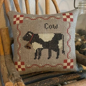 Peut inclure: Un oreiller gris avec des bordures à carreaux rouges et blancs présente une vache noire et blanche avec une cloche autour du cou. L'oreiller est brodé avec le mot "Cow".
