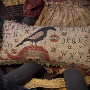 Peut inclure: Coussin décoratif rectangulaire avec un motif de corbeau. Le corbeau est noir et beige, tenant un ornement rouge. Le coussin a un motif d'alphabet et est bordé d'une frange verte. Le fond est de couleur beige.