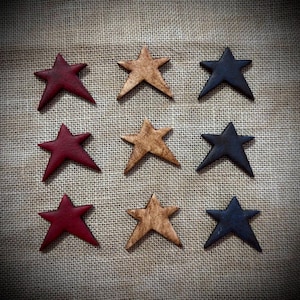 May include: Nine rustic star-shaped ornaments in a variety of colors. The stars are arranged in a 3x3 grid. Colors include deep red, tan, and navy blue. The stars are placed on a textured, neutral-colored fabric background.
