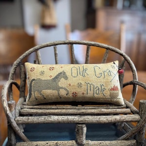 Peut inclure: Un coussin rectangulaire beige brodé avec les mots "Olde Gray Mare" en fil bleu foncé. Un cheval gris est brodé à gauche. Le coussin est orné d'accents floraux rouges et bleus et d'un petit charme de pomme rouge.